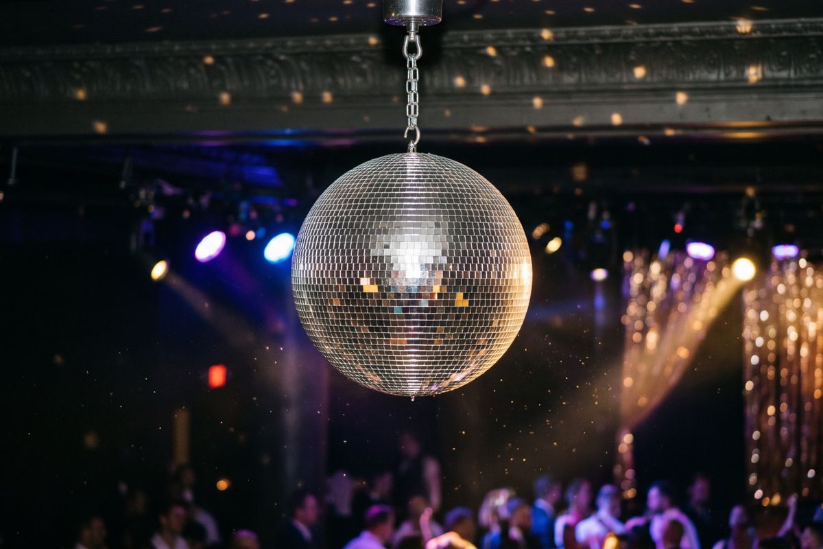 Mirrored disco ball hanging above a crowded dance floor, with purple and gold stage lights and a sparkling gold curtain behind