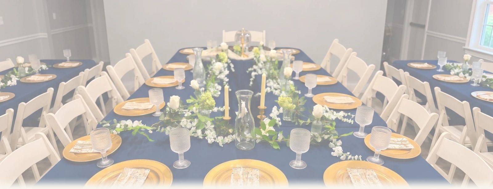 Long banquet table with a navy tablecloth, gold charger plates, patterned napkins, textured glass goblets, taper candles and a runner of white flowers and greenery, lined with white folding chairs